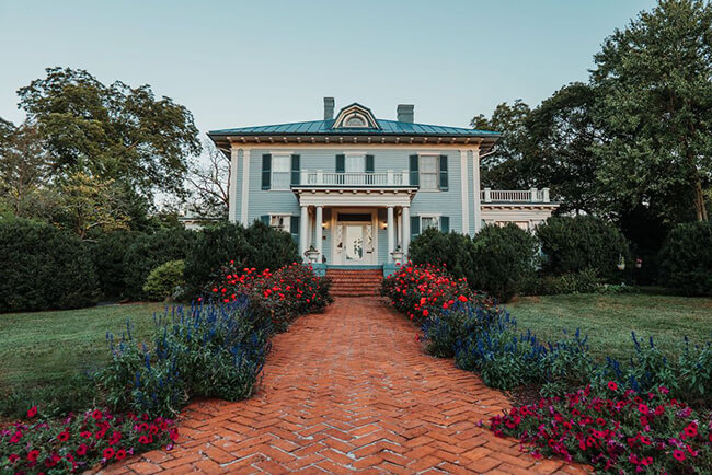A stately two-story colonial house with blue siding and a green roof, featuring a grand front porch supported by white columns. A vibrant brick walkway flanked by lush gardens of red and blue flowers leads up to the entrance. Green trees surround the property.