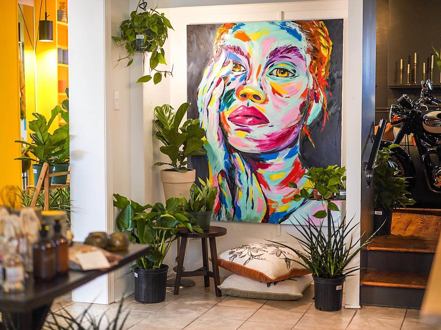 A vibrant painting of a woman's face, depicted in bold, multicolored brushstrokes, is displayed on a white wall. The room is filled with various green plants, adding a natural touch. A motorcycle is partially visible in the background, blending modern and artistic elements.