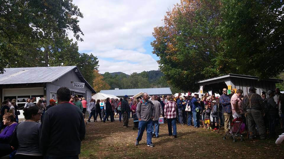 A bustling outdoor gathering with numerous people walking and socializing. Several small buildings and trees are in the background. A food stand is visible on the left. Many attendees are dressed in casual attire and the sky is partly cloudy.