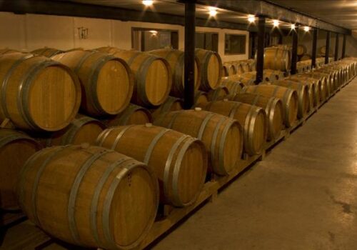 A dimly lit cellar with rows of wooden barrels stacked on wooden racks. The barrels are used for aging wine or spirits, and the cellar has a rustic ambiance with concrete floors and exposed beams on the ceiling. Image