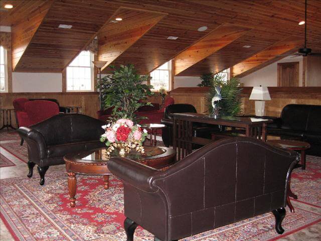 A spacious room with a vaulted wood ceiling features several dark leather sofas, red armchairs, and a central coffee table decorated with a floral arrangement. The floor is covered with patterned rugs, and various plants enhance the warm, inviting atmosphere.