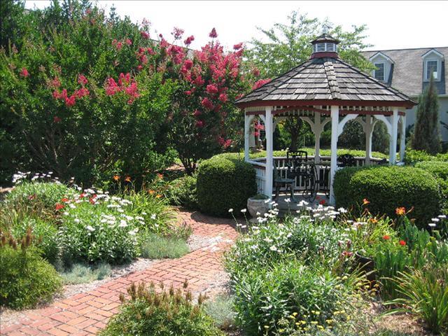 A picturesque garden features a white gazebo with a shingled roof, surrounded by lush greenery, blooming flowers, and manicured bushes. A brick path leading to the gazebo is flanked by tall trees and vibrant flower beds in full bloom. A house is visible in the background.