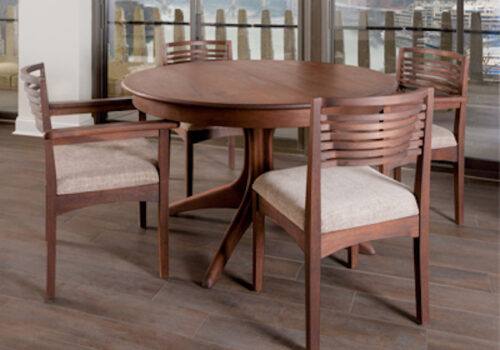 A round wooden dining table with four chairs, featuring cushioned seats and slatted backrests. The set is placed on a wooden floor near large windows, offering a scenic view of a waterfront with boats and buildings in the distance. Image