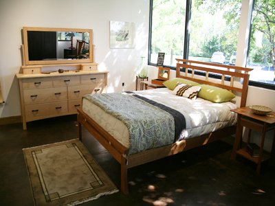 A cozy bedroom with a wooden bed frame, green and beige pillows, a patterned bedspread, a dresser with a mirror, and a nightstand. There is a rug in front of the bed, a painting on the wall, and large windows bringing in natural light.