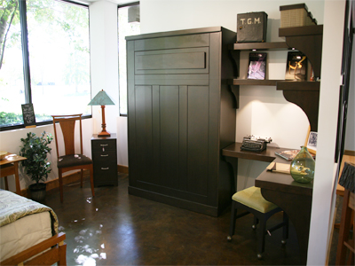 A compact, stylish home office features a dark wooden Murphy bed, a corner desk with shelving, and a chair. There are books, a typewriter, and decorative items on the desk and shelves. A window with greenery outside illuminates the space. A nightstand and chair are also present.