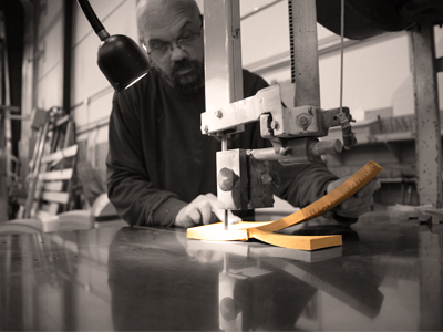 A person operates a band saw to cut a piece of wood in a workshop. The scene is lit by a task light positioned above the cutting area. The background shows equipment and shelves with various materials.