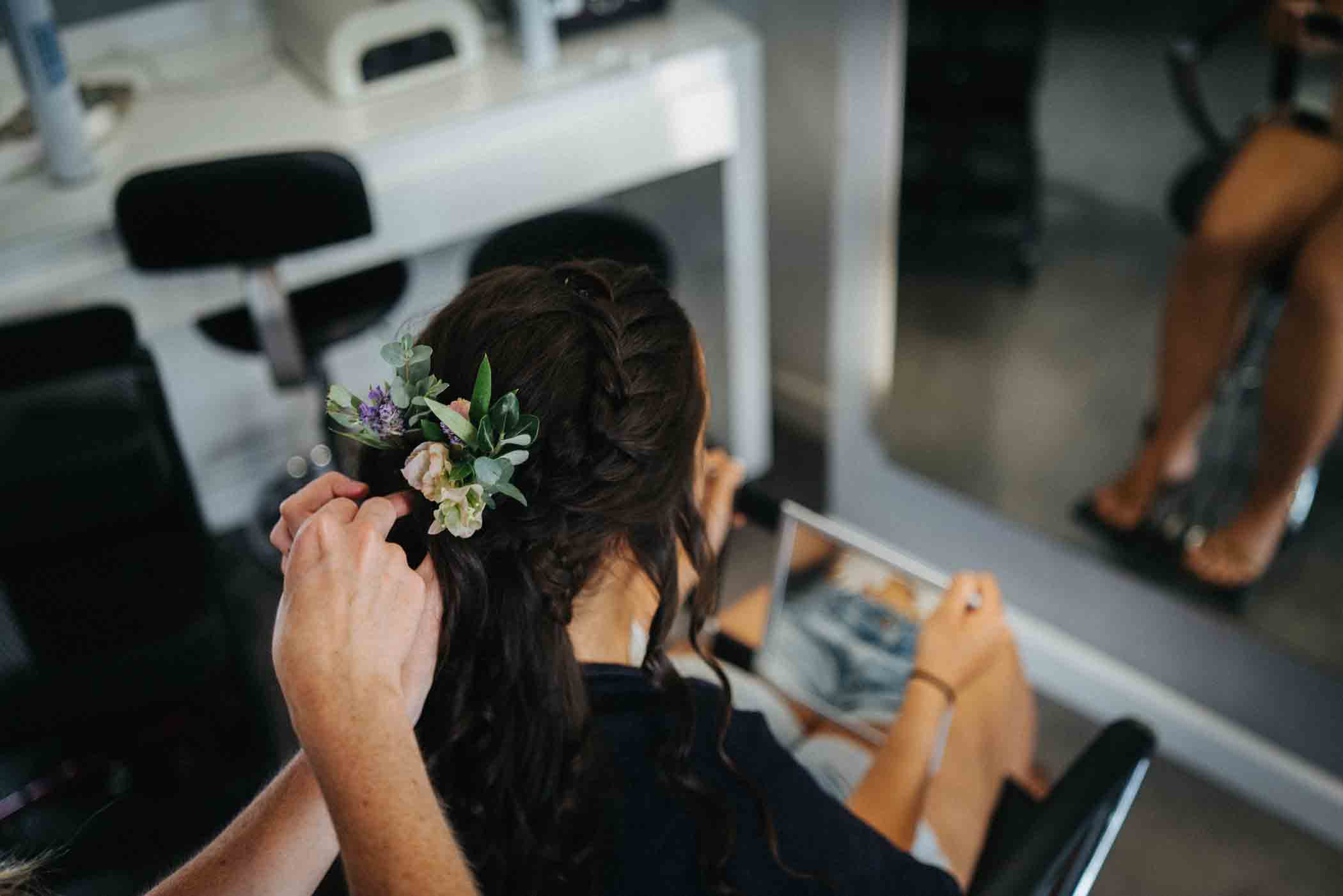 A person with long, dark hair is having flowers and greenery arranged in their braided hair by a stylist. They are sitting in a salon chair, holding a tablet or phone, with their reflection visible in a large mirror in front of them.