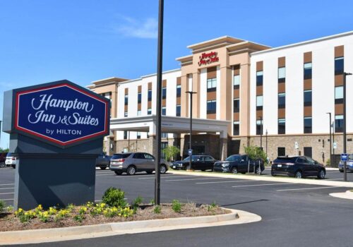 A Hampton Inn & Suites by Hilton hotel with a modern exterior featuring beige and white accents. The foreground shows the hotel's sign along with a parking lot containing several cars. The blue sky is clear and sunny. Image