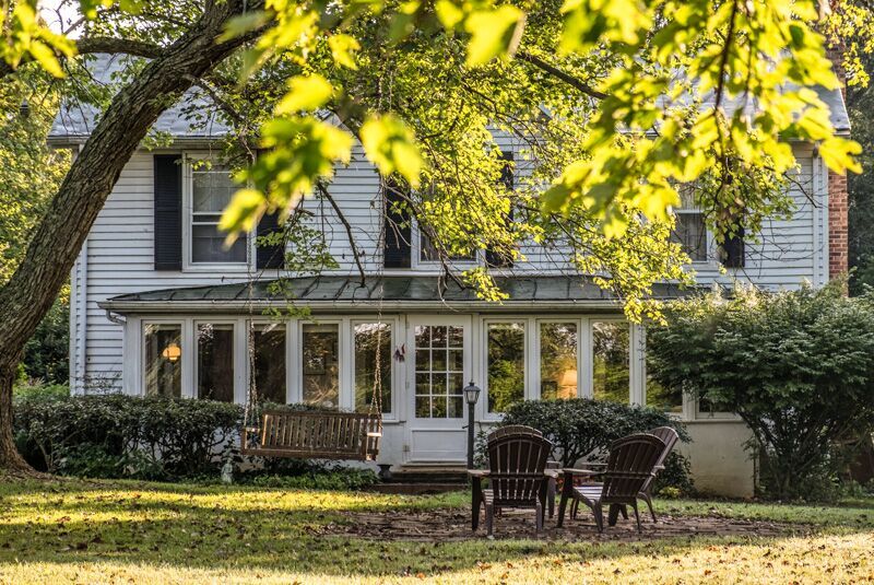 A charming white two-story house with a large front porch, surrounded by lush greenery. The yard features a wooden swing and two Adirondack chairs beneath a tree, suggesting a peaceful and relaxing outdoor space. The scene is bathed in warm sunlight.