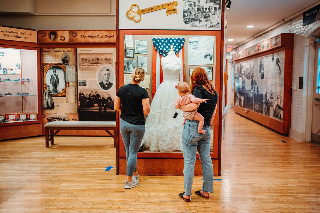 Two adults and a baby are viewing a white dress displayed in a glass case at a museum. The display is part of an exhibition with historical photographs and artifacts on the walls. The adults appear to be reading information about the dress.
