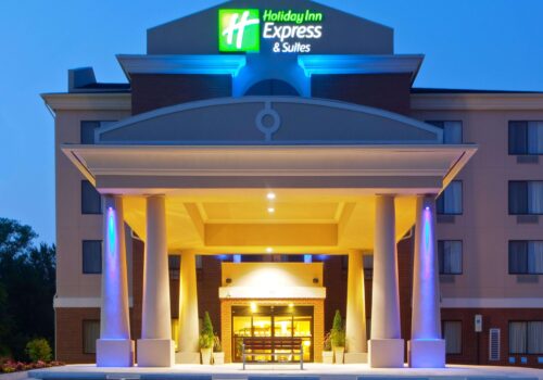 A front view of a Holiday Inn Express & Suites hotel building at dusk, showcasing its illuminated entrance with grand pillars and a modern sign on top of the structure. The building has multiple floors with lit windows, and the sky is beginning to darken. Image