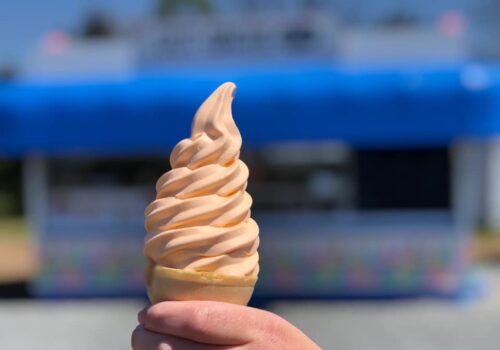 Close-up of a hand holding a cone with swirled, orange-tinted soft serve ice cream in front of a blurred blue and white building, likely an ice cream stand, on a sunny day. The background is out of focus, emphasizing the ice cream cone. Image