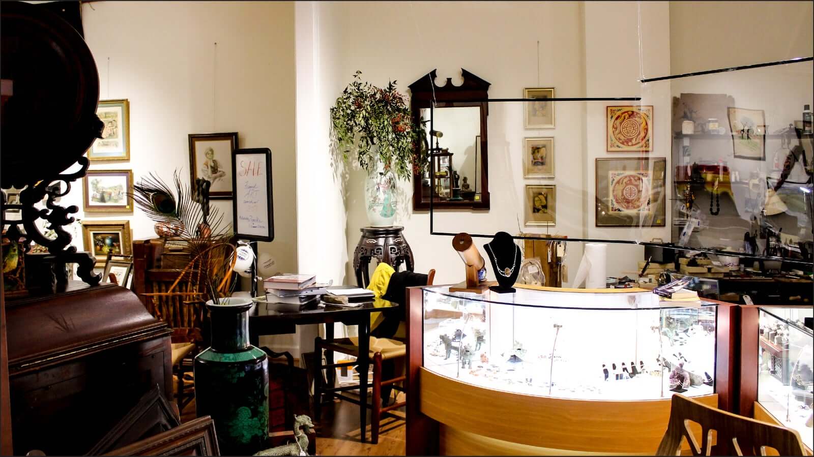 An antique shop with vintage furniture, paintings, and various decorative items on display. A glass counter showcases jewelry, and there's a round table with chairs filled with books and artifacts. The walls are adorned with framed pictures and greenery.