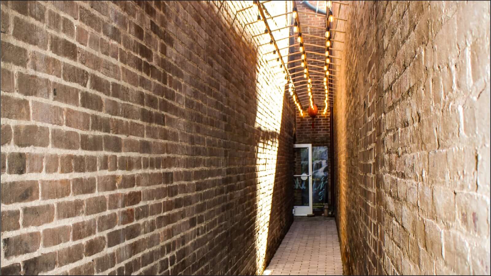 A narrow brick alleyway with a paved stone walkway stretches towards a glass door at the end. Sunlight filters through a lattice above, casting geometric shadows on the walls. The tall brick walls create a cozy, enclosed space.