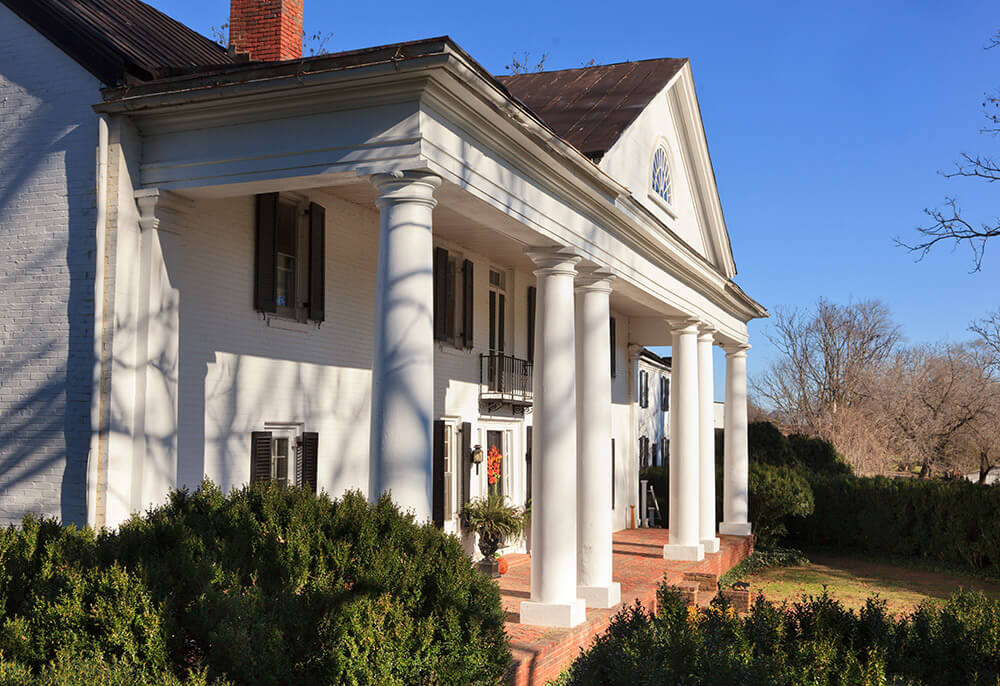 A large white colonial-style house with tall columns supporting a triangular pediment. The structure features a red brick staircase leading to the entrance and shuttered windows, surrounded by green bushes and trees, with a clear blue sky in the background.