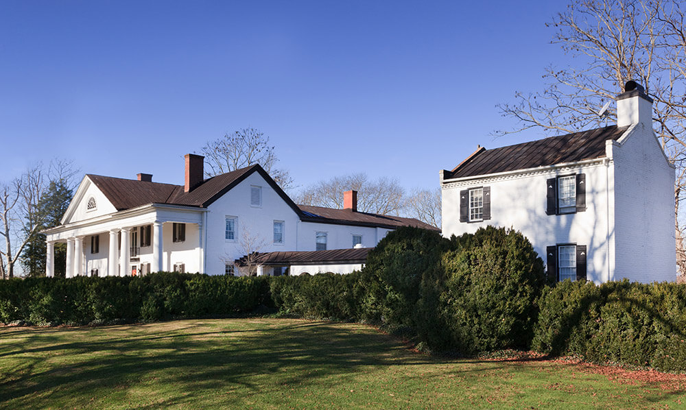 A large white colonial-style house with tall columns and black shutters is surrounded by a neatly trimmed hedge and green lawn. A smaller white brick building with a black roof is adjacent. The sky is clear and blue, and trees, some bare, are in the background.