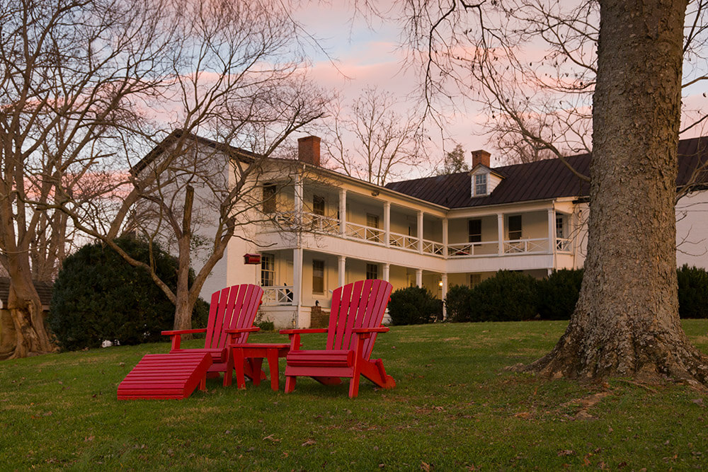 A picturesque two-story white house with wooden railings and a green lawn, set against a pink evening sky. Two red Adirondack chairs and a matching table are positioned under a tree in the foreground, surrounded by bare trees and bushes.