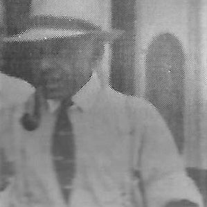 A grayscale image shows a man wearing a light-colored fedora, a shirt with rolled-up sleeves, a tie, and suspenders. He is smoking a pipe and looking down. The background appears to be a blurred doorway or window.