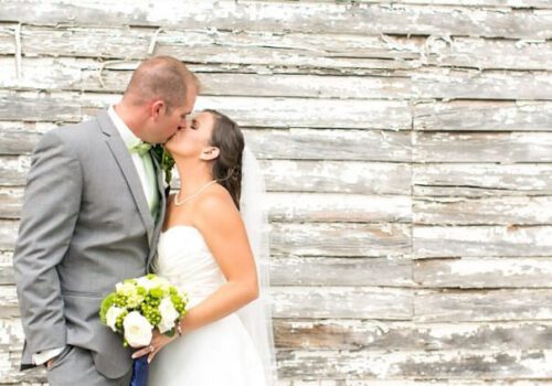 A bride in a white dress and veil kisses a groom dressed in a grey suit with a green tie. The bride holds a bouquet of white and green flowers. They stand in front of a weathered, rustic wooden wall. Image
