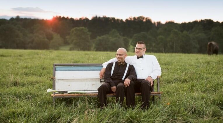 Two people are seated on an outdoor bench in a grassy field. One wears a black suit and bow tie, while the other wears a white shirt with a black bow tie and glasses. They sit closely, with one arm leaning on the other’s shoulder, in front of a backdrop of trees at sunset.
