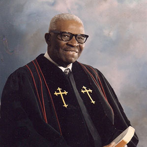 An older man with gray hair and glasses is wearing a black clerical robe adorned with gold crosses. He is holding a book and standing against a soft, cloudy background, smiling gently.
