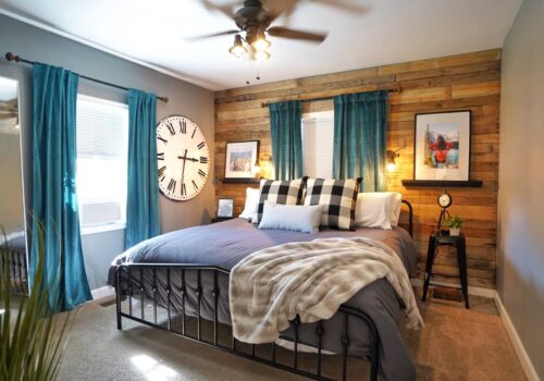 A cozy bedroom features a dark metal bed frame with gray bedding, plaid and white pillows, and a fur throw. The wood-accented wall behind the bed has two windows with blue curtains, while a large clock, framed pictures, and shelves decorate the space. Image
