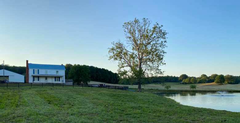 A serene countryside scene with a white farmhouse featuring a chimney, a blue roof, and a wraparound porch. The house is surrounded by a wooden fence, a large tree, and a pond reflecting the clear blue sky. The landscape is lush and green.