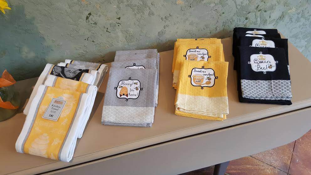 A display of neatly folded dish towels. The towels come in three color sets: white with yellow accents, gray with white designs, and black with yellow accents. Each set features fun, kitchen-related phrases and graphics on the front labels. They are placed on a taupe-colored table against a textured green wall.