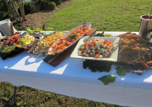 A buffet table set up outdoors on grass with a white tablecloth displays colorful platters of fresh fruit, a mixed salad with vegetables, a platter of cured meats, a bowl of fried foods, and a stack of plates at one end. The background shows greenery. Image