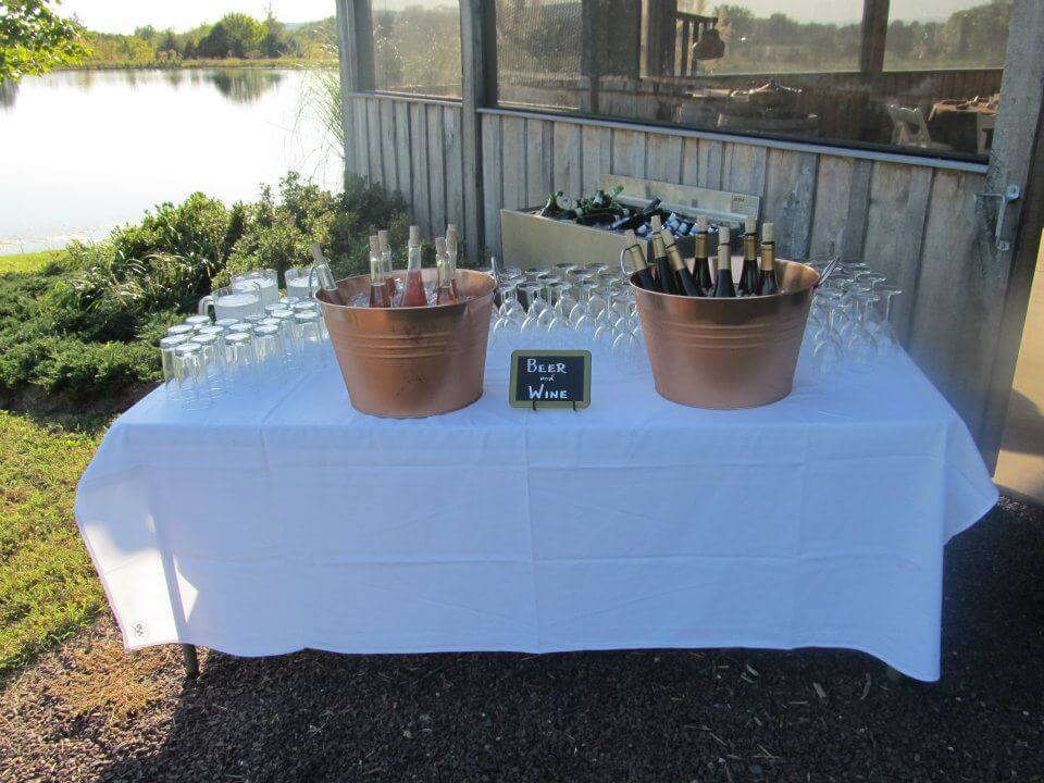 A table covered with a white cloth is set up outdoors beside a lake. On the table are two copper tubs, one with bottles of beer and the other with wine bottles. Wine glasses and a small sign labeled 