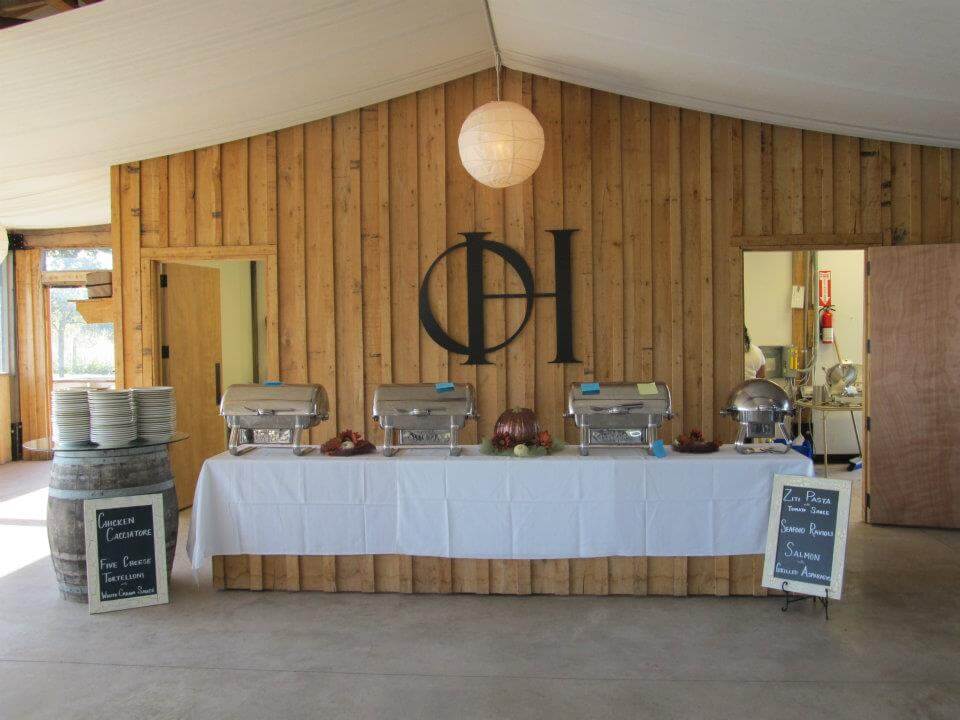 A rustic buffet setup with chafing dishes on a long table covered with a white tablecloth. To the left are stacked plates on a wooden barrel. Wooden walls in the background feature a circular light fixture and a large black circular logo. Two signs display menu items.