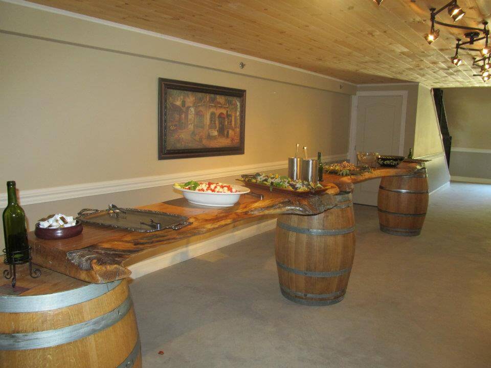 A rustic buffet setup with food placed on a long wooden table supported by wine barrels. Dishes include salads and appetizers. The table has wine bottles as decoration. The space has a wooden ceiling, soft lighting, and a painting on the back wall.