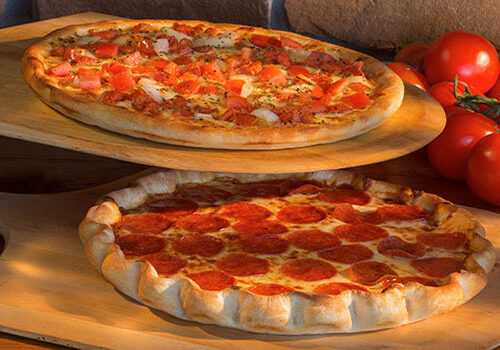 Two pizzas on wooden paddles. The top pizza is topped with diced tomatoes, onions, and herbs. The bottom pizza is a pepperoni pizza, with thick crust and a generous amount of pepperoni slices. Fresh tomatoes on the vine are in the background. Image