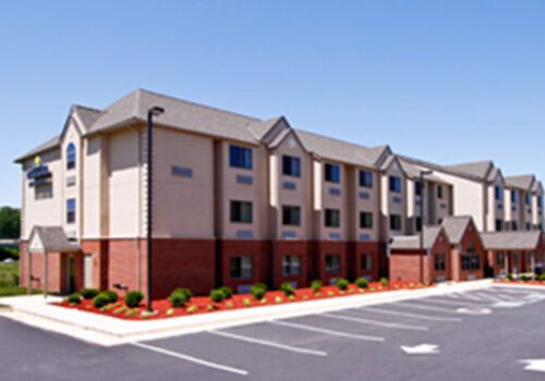 A three-story hotel with a red brick lower half and a beige upper half sits under a clear blue sky. The building features multiple windows and a landscaped area with shrubs along its base. The parking lot in front has several empty spots. Image