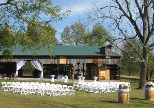 An outdoor wedding setup features rows of white chairs facing a rustic wooden barn with a green metal roof. The barn is decorated with white drapes, and two wooden barrels with white flowers stand at the entrance among the lush green grass and trees. Image