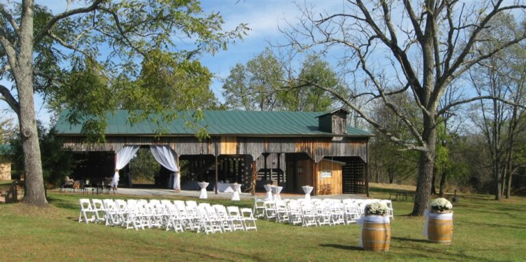 An outdoor wedding setup features rows of white chairs facing a rustic wooden barn with a green metal roof. The barn is decorated with white drapes, and two wooden barrels with white flowers stand at the entrance among the lush green grass and trees.