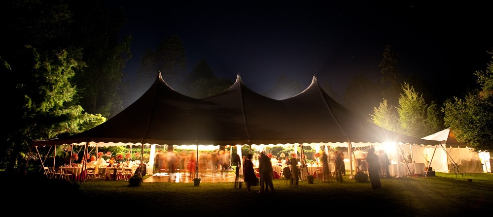 A large outdoor wedding reception at night under a tent with pointed peaks. Inside, tables and decorations are softly lit by warm lights. Guests, some blurred in motion, mingle and dance on a wooden dance floor. Trees surround the well-illuminated area.