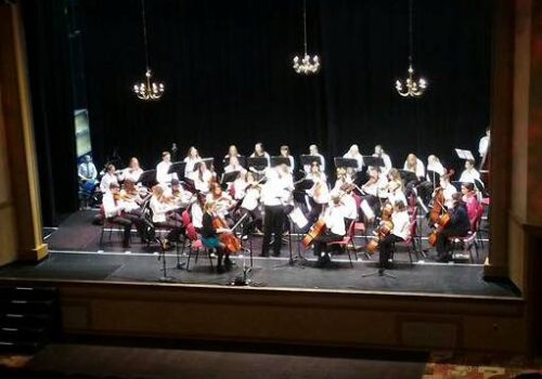 A group of musicians performs on stage in an orchestra setting. The conductor stands at the center, leading the ensemble. Various string, woodwind, and brass instruments are visible, with players seated in a semi-circle. The stage has black curtains and chandeliers above. Image