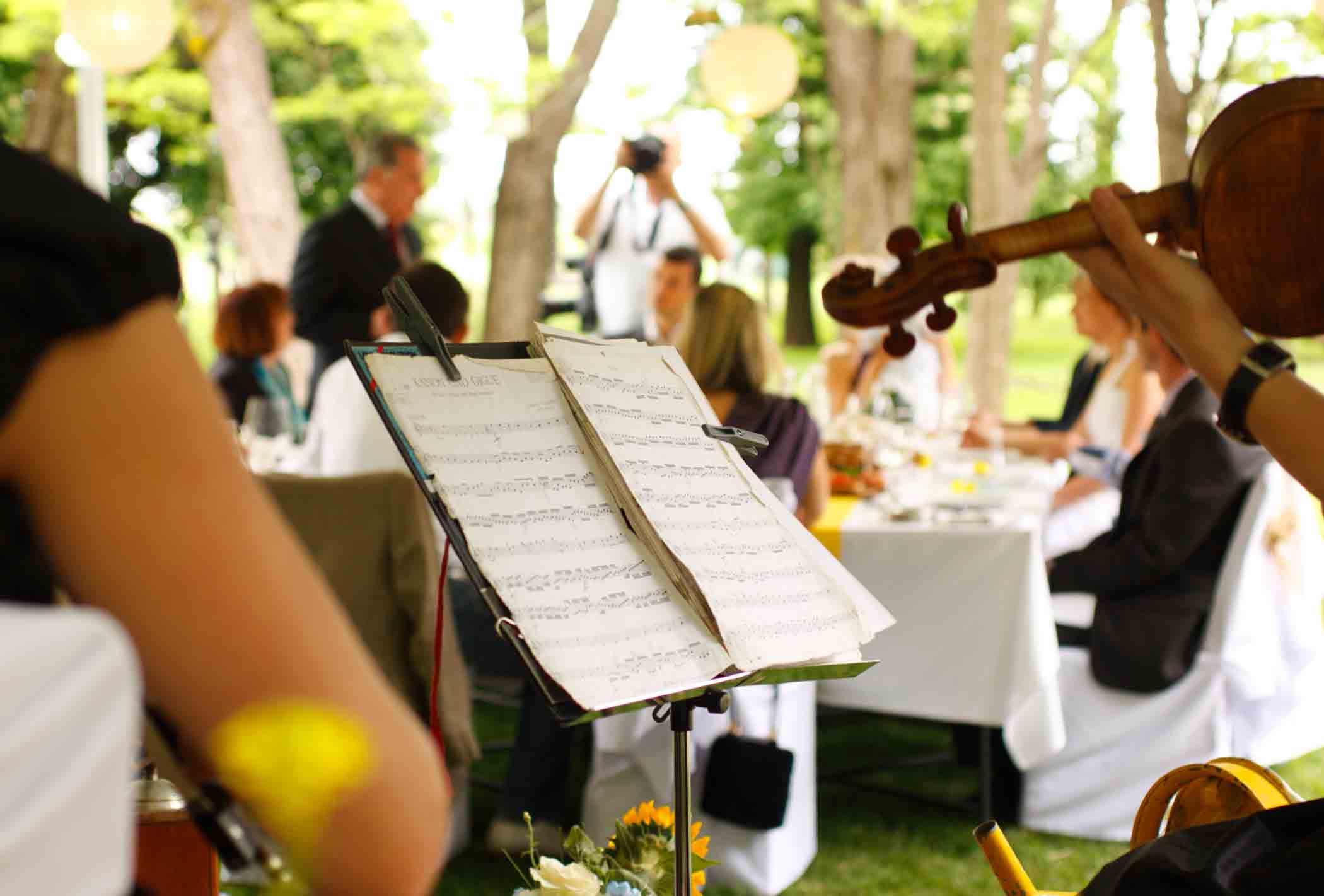 A close-up of a music stand with sheet music, surrounded by string instrument players. In the background, guests are seated at decorated tables outdoors, possibly attending a wedding or a formal event. Trees and greenery are visible beyond the tables.
