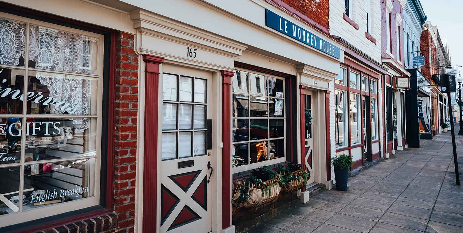A charming street with a row of small shops. One shop's window displays 'GIFTS' and has a vintage British style. Other shops have a mix of classic and modern facades, with door number 165 prominently visible. The scene is quiet and inviting.