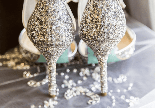 A pair of glittery high-heeled shoes with silver sparkles is placed on a delicate, lace-like fabric with floral embroidery. The shoes have ankle straps and pointed toes, with the heels facing the viewer. The background features soft, diffused lighting. Image