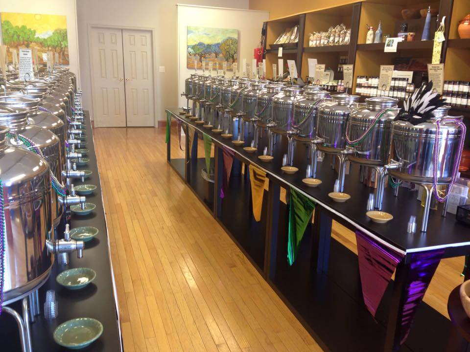 A store interior with rows of shiny stainless steel containers on black shelves, likely for oil or vinegar. Each container has a spout and a small dish underneath. The walls display colorful artwork, and there are shelves with various items at the back.
