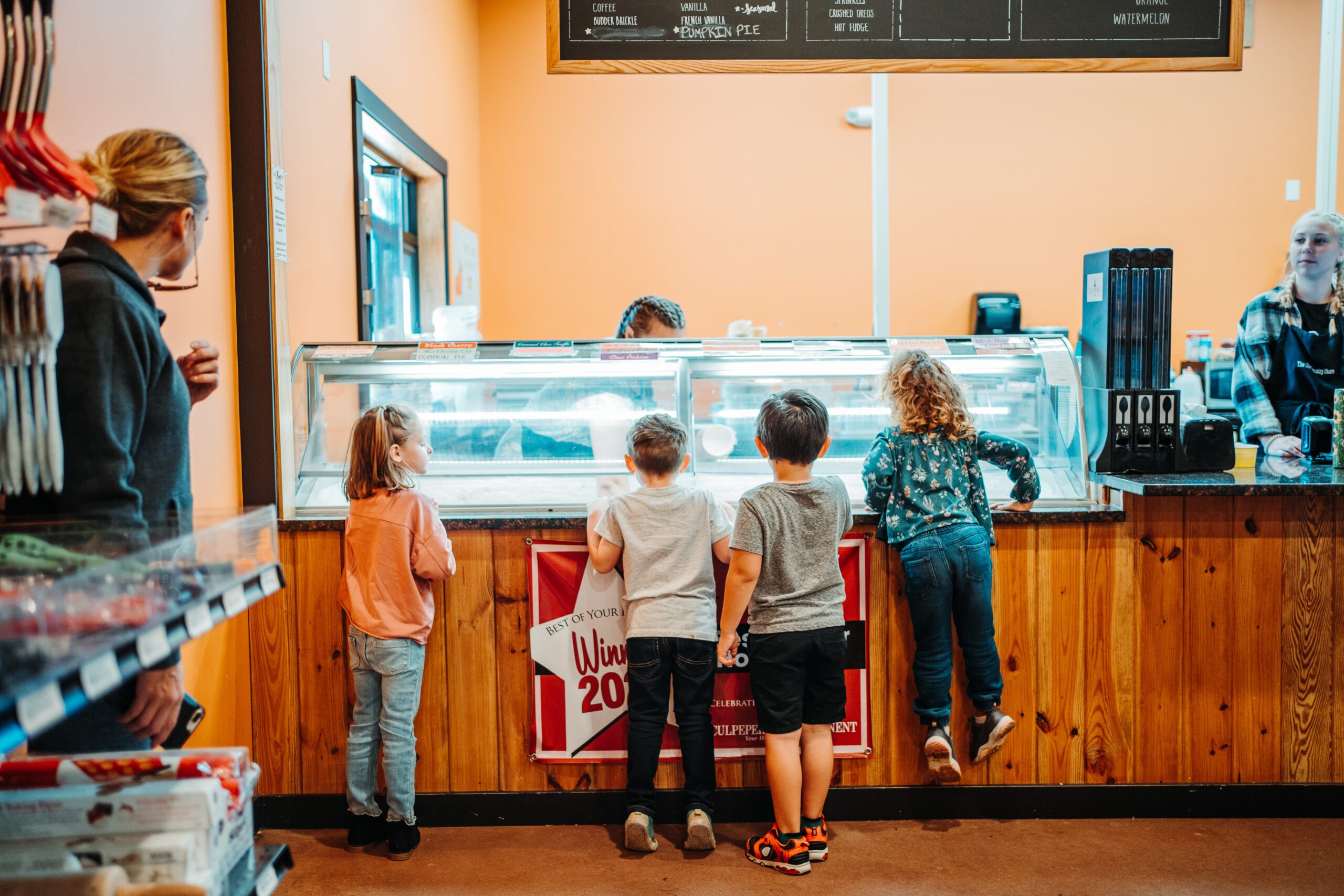 Five children stand in front of a display case at an ice cream shop, eagerly looking at the options. An adult stands nearby, and a staff member behind the counter is ready to serve them. The shop has a cozy, welcoming atmosphere with orange walls and wooden accents.