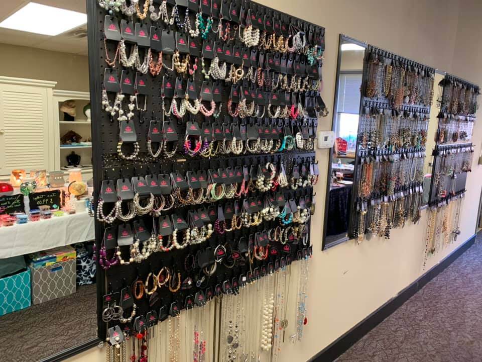 A wall display showcases a variety of hanging jewelry, including necklaces and earrings. Each piece is meticulously arranged on black boards. To the left, a table with colorful accessories and decor is partially visible. The setting appears to be a boutique or small shop.