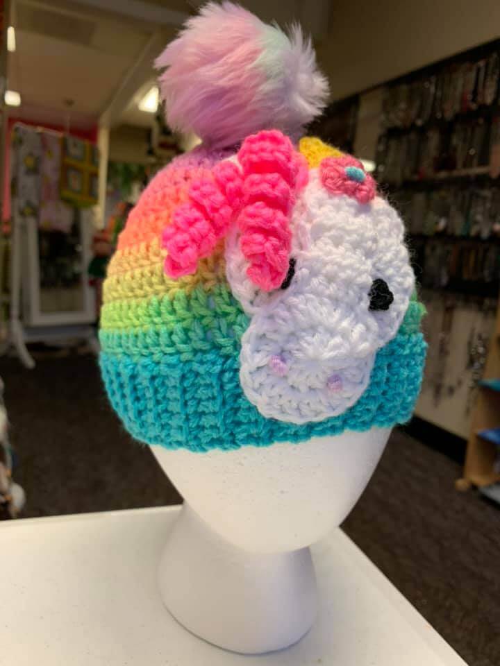 A white mannequin head displays a colorful, hand-knitted hat with a fuzzy pom-pom on top. The hat features a rainbow gradient pattern and a crocheted unicorn face with a pink mane on the front. The background includes shelves and displays of various items.
