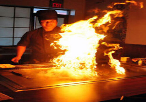 A chef wearing a dark uniform and hat performs a cooking demonstration at a teppanyaki grill, with large flames erupting from the grill in front of them. The kitchen's dim lighting emphasizes the bright fire. Image