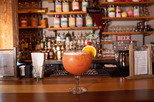 A colorful tropical cocktail garnished with a mini umbrella, orange slice, and cherry sits on a wooden bar counter. Behind the drink are liquor bottles and rows of colorful cylindrical containers on shelves, creating a lively and vibrant bar atmosphere.