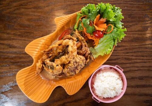 A dish of crispy fried soft-shell crab is artfully presented on a fish-shaped plate, garnished with fresh vegetables including lettuce, carrot, and cherry tomatoes. A bowl of white rice accompanies the meal, all set on a wooden surface. Image
