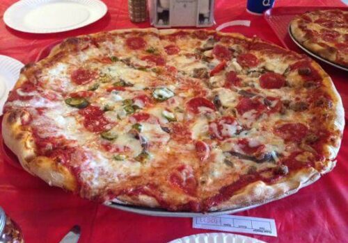 A large pizza topped with a variety of ingredients such as pepperoni, mushrooms, green peppers, and cheese sits on a red tablecloth. There are paper plates, a pepper shaker, and a blue Pepsi can in the background. Image