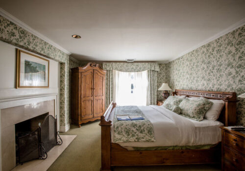 A cozy bedroom featuring a wooden bed with white and green floral bedding, matching the floral wallpaper. There is a wooden armoire beside a window with sheer curtains letting in natural light. A white fireplace with a screen is on the left wall, with a painting above it. Image